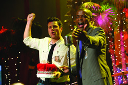 A man in a white shirt raises his fist while a man in a grey suit gestures toward the audience.