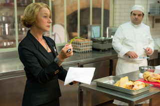 A woman in a black blazer holds a green vegetable while a chef stands behind a stainless steel counter.
