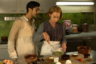 A man in a cream tunic watches a woman in a plaid shirt mix ingredients in a glass bowl.