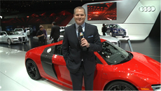A man in a dark suit stands in front of a red Audi sports car at an indoor automotive exhibition.