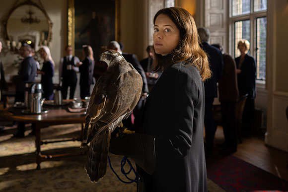 A woman in a black coat holds a large hawk while standing in a room with people in the background.