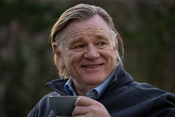 A smiling middle-aged man with light hair holding a dark cup in an outdoor setting.