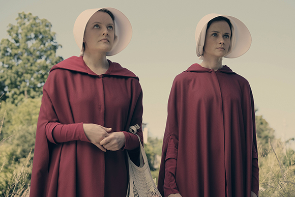 Two women wearing long red capes and large white bonnet-style headpieces standing in a field.