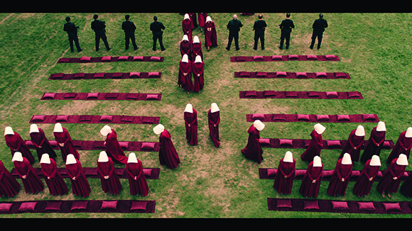 People in black suits and red hooded robes arranged in symmetrical rows on a green field.