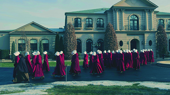 A long line of people wearing red robes and white head coverings walking in front of a large stone mansion.