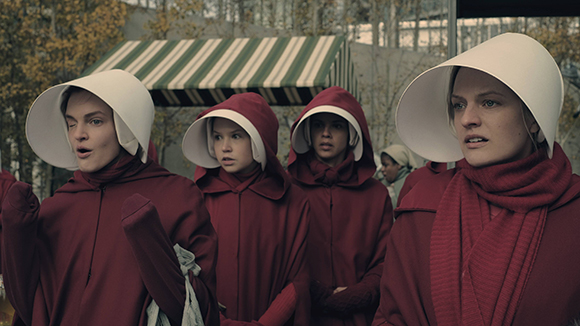 Four women wearing matching red hooded robes and large white bonnet-style headpieces.