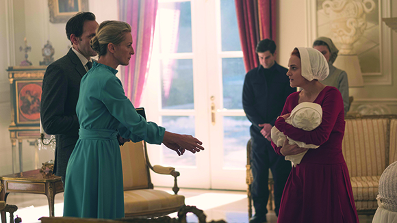 A woman in a teal dress gestures toward a woman in a red dress holding a baby.