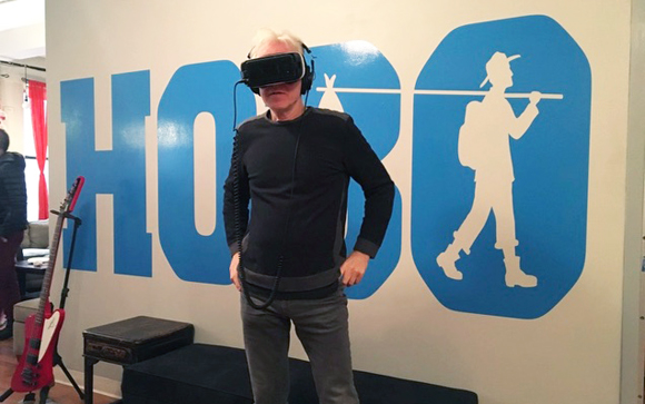 A man wearing a virtual reality headset stands before a large blue logo that reads 'HOBO' with a silhouette icon.