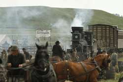 A steam train emits smoke near horse-drawn wagons and people in a historical frontier town.