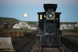A black steam locomotive on tracks facing forward with a full moon in the twilight sky behind it.