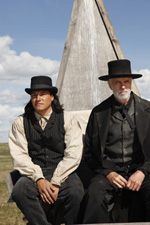 Two men in 19th-century clothing and black hats sit in front of a tall wooden structure under a blue sky.