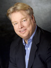 A smiling man with blond hair wearing a dark blazer and a light blue button-down shirt.