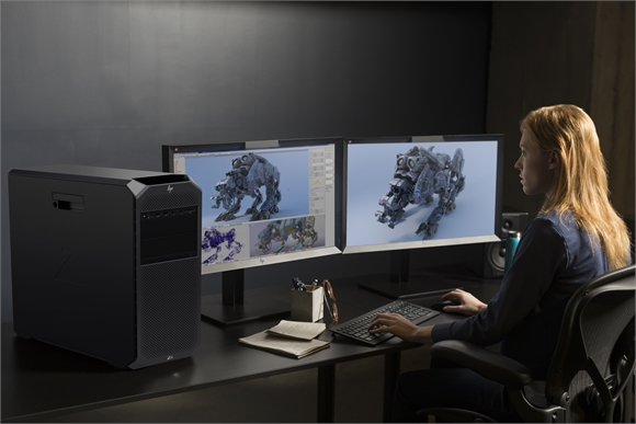 A woman sits at a desk using two monitors displaying 3D robot models next to a large black computer tower.