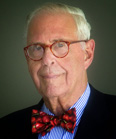 An elderly man with white hair, red-rimmed glasses, and a red patterned bowtie wearing a blue striped shirt.