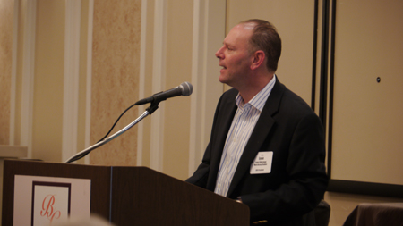 A man in a dark blazer and striped shirt speaks into a microphone at a podium.