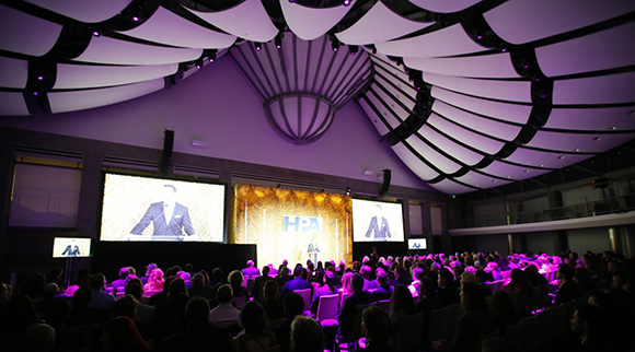Audience seated in a dark auditorium watching a presentation on large screens under a curved, patterned ceiling.