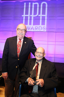 A tall man in a navy blazer and red tie stands next to a seated man in a dark suit holding an award.