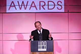 A man in a dark suit gestures while speaking at a podium with HPA Awards branding against a pink background.