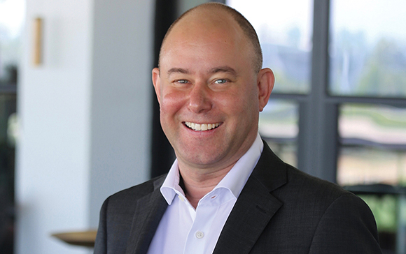 A smiling man with a shaved head wearing a dark blazer and a white button-down shirt.