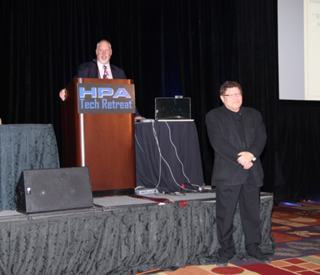 A man in a suit speaks at a podium with the HPA Tech Retreat logo. Another man in a suit stands to his right on a stage.