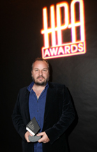 A man in a dark velvet blazer holding a small award in front of a glowing HPA Awards neon sign.
