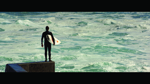A person in a black wetsuit stands on a concrete pier holding a surfboard, facing choppy turquoise ocean waves.