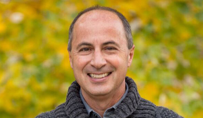 A middle-aged man with short dark hair and a grey sweater smiling against a blurred yellow background.