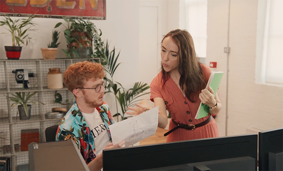 A woman in a red dress holds a green folder and points to a paper held by a man in a floral shirt.