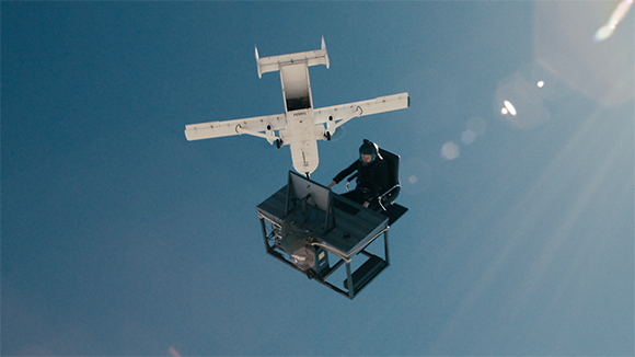 A white fixed-wing drone flies above a person sitting at a mobile control station against a clear blue sky.