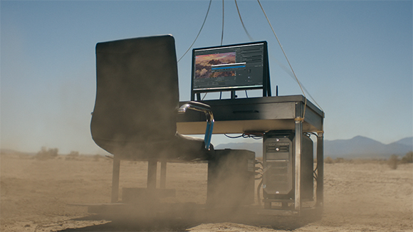 A desk, chair, monitor, and computer tower suspended by wires over a dusty desert ground.