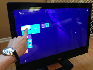 A person's hand reaches out to touch a blue Windows Start screen on a large monitor.