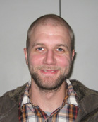 A man with short hair and a beard wearing a plaid shirt and a jacket.