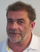 A man with short, messy brown and grey hair and a matching beard wearing a white polo shirt.