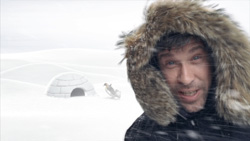 A man wearing a fur-lined hood smiles in a snowy landscape with an igloo behind him.