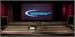 A large audio mixing console in a dark studio with a screen displaying a blue logo in the background.