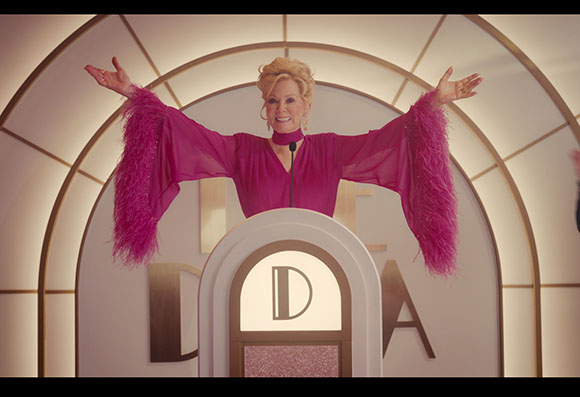 A woman in a bright pink dress with large feathered sleeves stands behind a podium with arms raised.