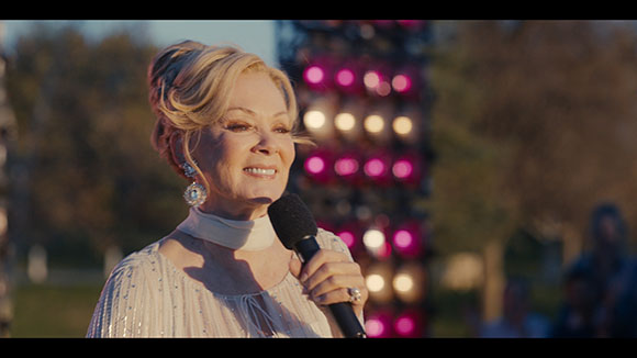 A blonde woman in a white sequined top smiles while holding a black microphone outdoors.