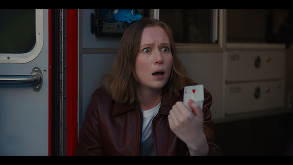 A woman with light brown hair wearing a brown leather jacket looks surprised while holding two playing cards.