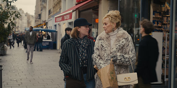 Two women stand on a city sidewalk talking, one in a dark cap and striped jacket, the other in a white fur coat.