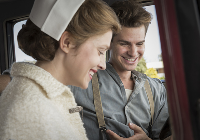 A smiling woman in a white nurse's cap and a man in a grey shirt and suspenders look down together.