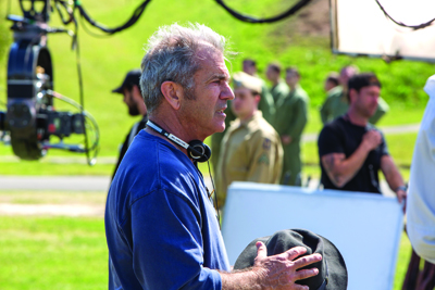 A man with gray hair in a blue shirt stands outdoors on a film set with crew and equipment in the background.