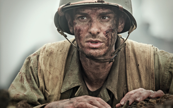 A soldier in a helmet and tan uniform with mud on his face looks forward with a focused expression.