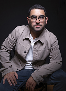 A man with dark hair and glasses wearing a tan jacket and white shirt sits against a black background.