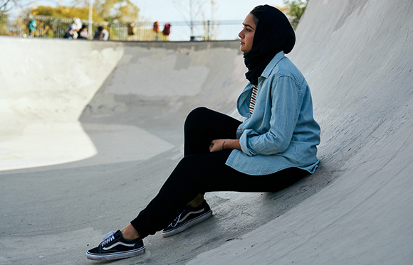 A woman in a black hijab, blue denim shirt, and black pants sits on the edge of a concrete skatepark bowl.