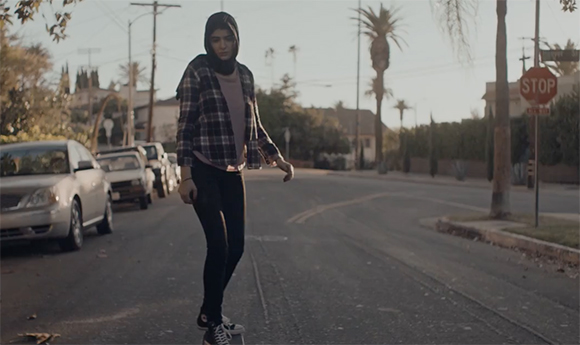 A person wearing a hijab, plaid shirt, and black leggings rides a skateboard down a quiet street.