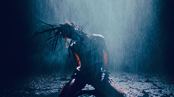 A silhouette of a person with long hair dancing in heavy rain under a bright blue spotlight.