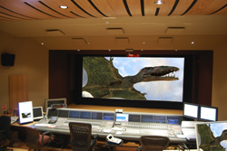 A large screen in a dark studio shows a CGI dinosaur, positioned above a wide sound mixing console.