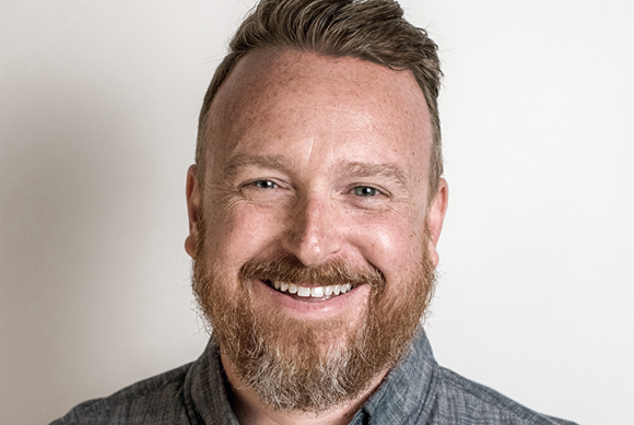 A smiling man with reddish-brown hair and a full beard against a white background.