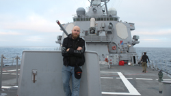 A man in a black hoodie and jeans leans against a gray metal structure on a ship deck at sea.