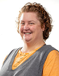 A woman with curly brown hair and a gray vest over an orange shirt smiles.